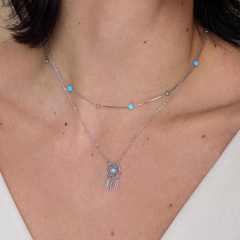 May include: A layered silver-tone necklace featuring two delicate chains. The upper chain is adorned with small, round, light blue stones, while the lower chain holds a circular dreamcatcher pendant with a matching blue stone and three small feathers.