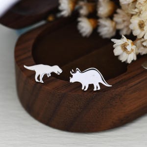 Op de afbeelding: Zilveren dino-oorstekers. Eén oorbel heeft een Tyrannosaurus Rex, de andere een Triceratops. De oorbellen worden gepresenteerd op een donker houten oppervlak met witte bloemen. Een uniek sieradengeschenk.