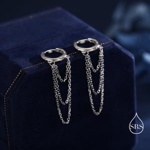 Może przedstawiać: Para srebrnych kolczyków koła z wiszącymi łańcuszkami. Kolczyki są prezentowane na ciemnoniebieskim aksamitnym tle. Łańcuszki zwisają warstwowo, dodając ruch i wizualne zainteresowanie biżutrą.