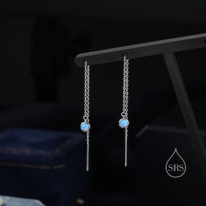 Puede incluir: Un par de pendientes de plata con una piedra de ópalo azul claro en cada pendiente. Los pendientes tienen una cadena delicada y una fina varilla de plata que cuelga de la piedra.