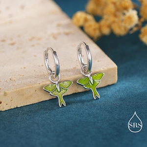 May include: A pair of silver hoop earrings with green moth pendants. The moth pendants have a detailed design with green enamel and silver outlines. The earrings are displayed on a neutral-colored surface against a dark teal background.