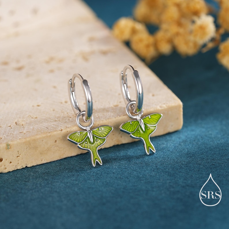 May include: A pair of silver hoop earrings with green moth pendants. The moth pendants have a detailed design with green enamel and silver outlines. The earrings are displayed on a neutral-colored surface against a dark teal background.