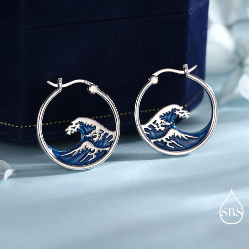 Puede incluir: Un par de pendientes de aro de plata con un dise&ntilde;o circular y una incrustaci&oacute;n de esmalte que representa una ola oce&aacute;nica estilizada en azul y blanco. Los pendientes tienen un mecanismo de cierre de bisagra y un sello 925 en el aro.