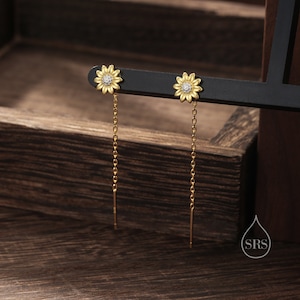 Puede incluir: Un par de pendientes colgantes de oro con un diseño de girasol y un solo cristal transparente en el centro de cada flor. Los pendientes cuelgan de una cadena de oro.