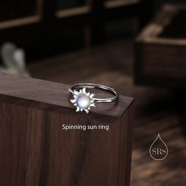 Puede incluir: Un anillo de plata con un dise&ntilde;o de sol de piedra lunar. El anillo est&aacute; sobre una superficie de madera con el texto "Spinning sun ring" debajo.