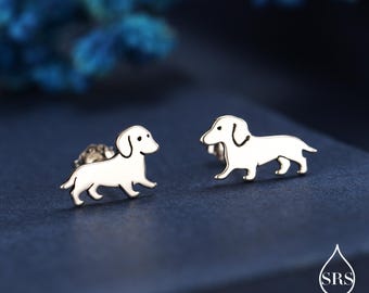 Op de afbeelding: Een paar zilverkleurige teckel oorknopjes. Elke oorbel heeft een gestileerd honden silhouet met een gebogen staart en een kleine, ronde kop. De oorbellen zijn geplaatst tegen een donkerblauwe achtergrond.