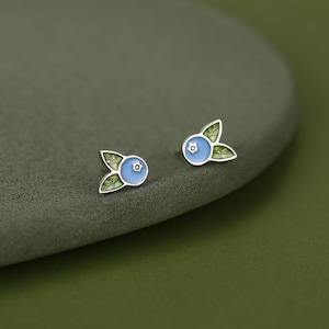 Op de afbeelding: Een paar oorknopjes in de vorm van bosbessen, voorzien van een lichtblauw cirkelvormig midden met een klein vijfpuntssterdetail, geaccentueerd door twee groene blaadjes met zichtbare nerven, gevat in een gepolijst zilverkleurig metalen frame.