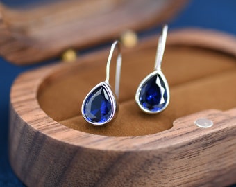 May include: A pair of silver teardrop earrings with blue gemstone accents. The earrings are displayed in a wooden box.