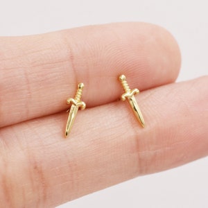 Könnte beinhalten: Ein Paar goldene Dolch-Ohrringe. Die Ohrringe sind klein und zart und haben ein schlichtes, klassisches Design.