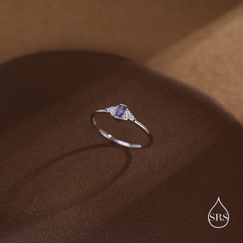 Puede incluir: Un delicado anillo de plata con una peque&ntilde;a piedra preciosa rectangular de color p&uacute;rpura rodeada de piedras preciosas m&aacute;s peque&ntilde;as de color claro.