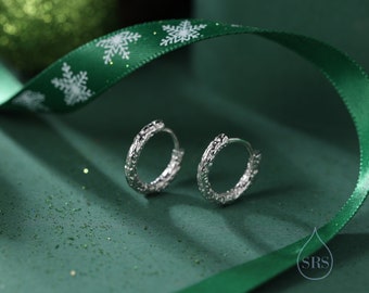 May include: A pair of silver hoop earrings with a textured design. The earrings are on a green background with a green ribbon with white snowflakes.