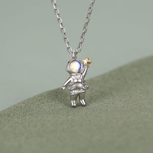 May include: A silver-coloured necklace featuring a pendant shaped like an astronaut. The astronaut figure has a round, iridescent stone for a helmet visor and is reaching up to touch a small, gold-coloured star held in one hand. The chain is a simple link style.