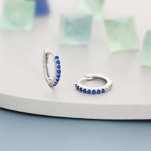 Puede incluir: Un par de pendientes de aro de plata con una fila de piedras preciosas azules alrededor del borde.