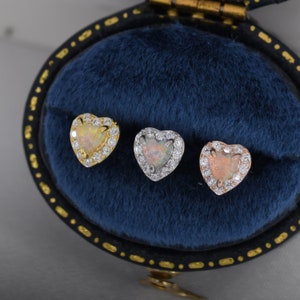 May include: Three heart-shaped opal earrings with a halo of clear cubic zirconia stones. The earrings are displayed in a blue velvet box. The earrings are available in gold, silver, and rose gold.