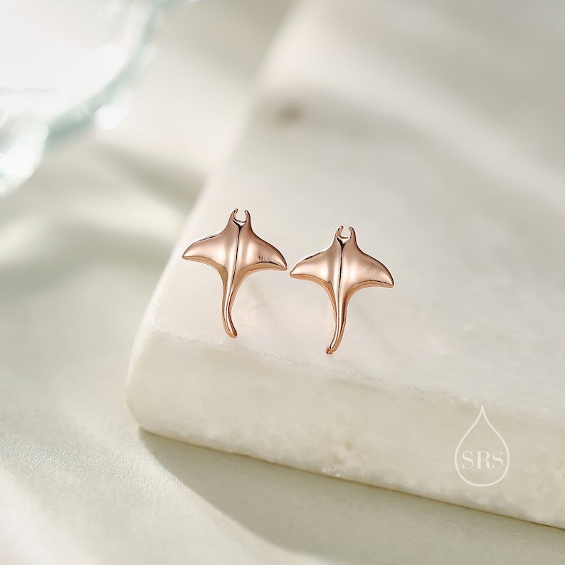 May include: A pair of rose gold stud earrings featuring a manta ray design.