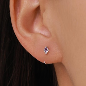 May include: A silver stud earring with a purple gemstone in a diamond shape is shown in a close-up. The earring is in an ear. A small silver hoop earring is also visible.