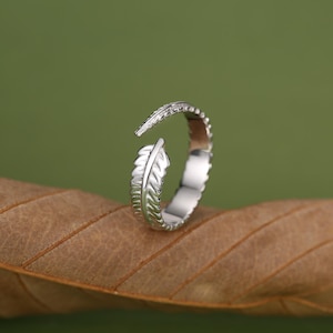 May include: A silver-toned open ring featuring a detailed feather design that wraps around the finger. The band has a polished metallic finish with etched lines mimicking the texture of a feather, with the two ends meeting in the centre.