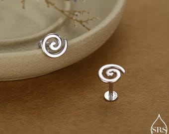 Op de afbeelding: Een paar zilverkleurige spiraalvormige oorstekers. Eén oorbel wordt op een plat oppervlak getoond en de andere apart. De oorbellen hebben een minimalistisch ontwerp, met een wervelvorm. De achtergrond is een warme, neutrale tint.