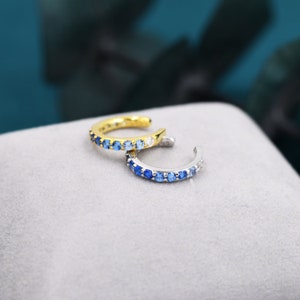 May include: Two ear cuffs with a gradient of blue gemstones. The ear cuff on the left is gold and the ear cuff on the right is silver.