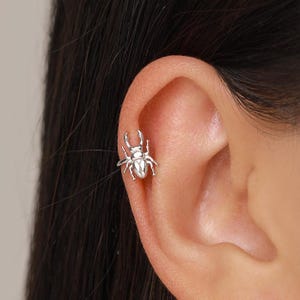 May include: A silver-toned ear cuff shaped like a stag beetle. The jewelry piece features a polished metallic finish with detailed legs and mandibles, designed to wrap around the outer edge of the ear cartilage for a secure fit.