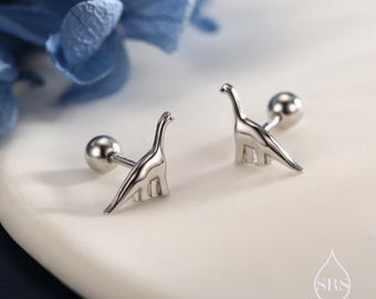 May include: A pair of silver-colored dinosaur stud earrings. Each earring features a stylized dinosaur silhouette with a rounded post and ball backing. The earrings are displayed on a white surface with a blurred blue floral background.