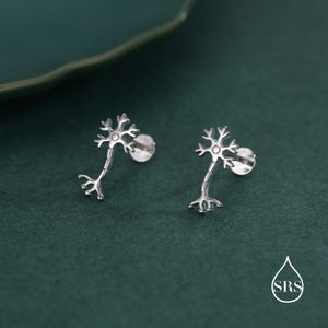 May include: A pair of silver-colored stud earrings shaped like neurons. Each earring features a central cell body with radiating dendrites and an elongated axon. The earrings are set against a dark green background.
