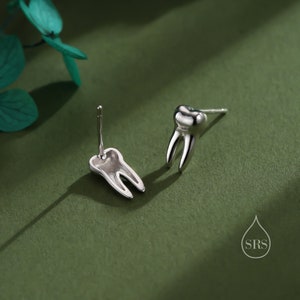 May include: A pair of silver-toned stud earrings shaped like human molars. Each earring features a polished, metallic finish with a distinct root structure and a post backing. A small logo with a water drop shape and the letters SRS is visible.