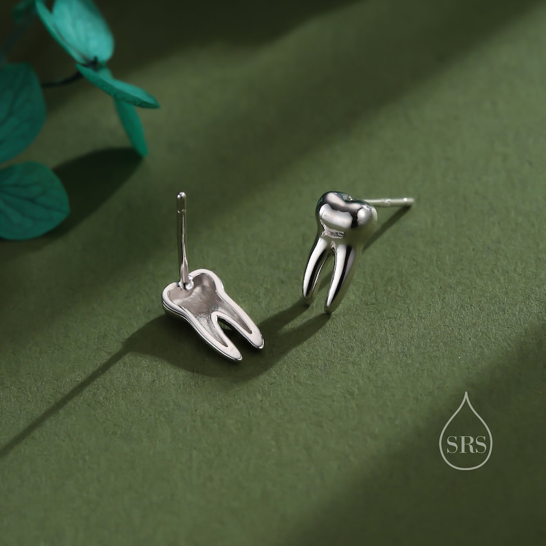 May include: A pair of silver-toned stud earrings shaped like human molars. Each earring features a polished, metallic finish with a distinct root structure and a post backing. A small logo with a water drop shape and the letters SRS is visible.