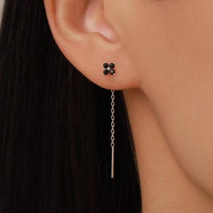 Pode incluir: Um brinco de corrente de prata com um pino em forma de flor de quatro pétalas cravejado com quatro pequenas pedras pretas. O brinco inclui uma delicada corrente de prata que pende do pino, terminando numa barra de metal reta e fina.