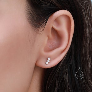 May include: A close-up of a person's ear with a small silver earring with three clear round stones in a curved line.