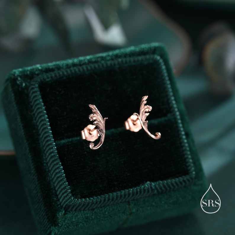 May include: A pair of rose gold earrings with a delicate, leaf-shaped design. The earrings are displayed in a green velvet box.