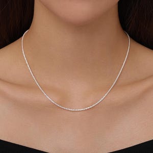 Op de afbeelding: Een dunne, glinsterende zilveren ketting met een getextureerd, gedraaid touwontwerp. De metalen schakels vangen het licht op en creëren een helder, reflecterend oppervlak dat in een soepele bocht rond de nek ligt voor een eenvoudige uitstraling.