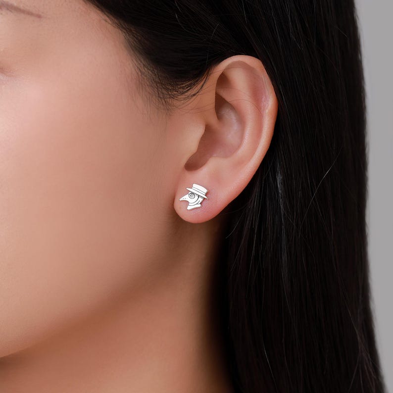 May include: A pair of silver-colored stud earrings. Each earring features a detailed design of a plague doctor's mask and hat. The earrings are small and delicate, suitable for everyday wear. The earrings are shown on an ear.