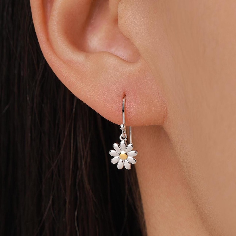 May include: A silver daisy earring with a gold center is shown hanging from an ear. The earring has a hook closure. The daisy petals are white. The earring is a delicate piece of jewelry.