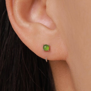 May include: A close-up of an ear with a small, round, light green stud earring. The earring is set in a silver-colored metal. A small silver hoop earring is below the stud.