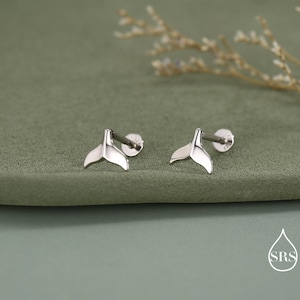 May include: A pair of silver stud earrings in the shape of whale tails. The earrings are presented on a green surface, with a sprig of dried flowers in the background. Each earring has a post and a small, round backing.