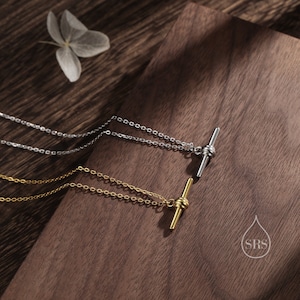 May include: Two delicate necklaces with a simple bar pendant featuring a knotted design. The necklace on top is silver, and the necklace on the bottom is gold. Both necklaces are displayed on a wooden surface.