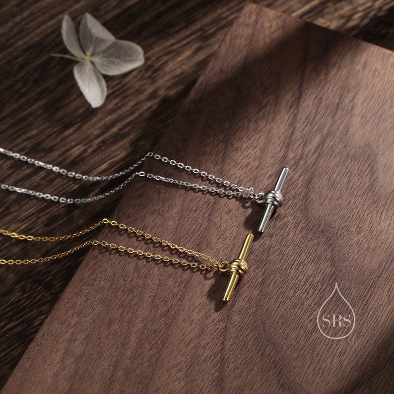 May include: Two delicate necklaces with a simple bar pendant featuring a knotted design. The necklace on top is silver, and the necklace on the bottom is gold. Both necklaces are displayed on a wooden surface.