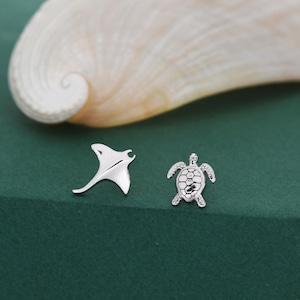 May include: Silver-toned stud earrings featuring a manta ray and a sea turtle design. The earrings are displayed on a dark green surface with a seashell in the background. The manta ray earring is a stylized shape, and the turtle earring has a detailed shell.