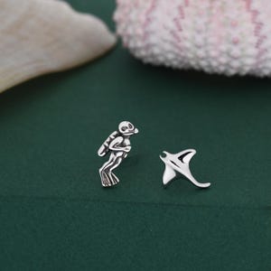 May include: Silver-toned diver and manta ray earrings. The diver is depicted with a diving mask and air tank. The manta ray has a curved, wing-like shape. The earrings are set against a dark green background.