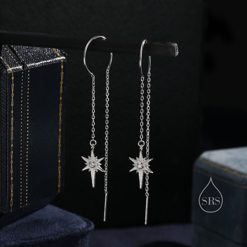 May include: A pair of silver dangle earrings with a star-shaped charm set with clear cubic zirconia stones. The earrings have a long chain that hangs down from the star.