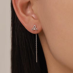 May include: A silver-colored earring set featuring a Celtic knot stud and a chain with a bar drop. The earring is displayed on an ear, showcasing the design and length of the chain. The jewelry is simple and elegant.