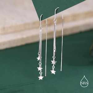 May include: A pair of silver dangle earrings with three star charms hanging from each chain. The earrings are on a green background.