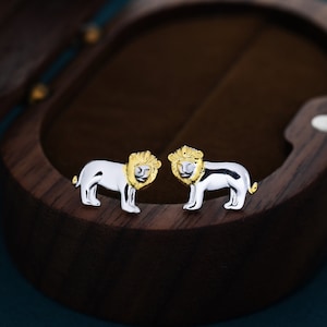May include: A pair of gold and silver lion stud earrings. The lions are facing forward with their manes and faces in gold and their bodies in silver.