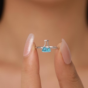 Könnte beinhalten: Ein silberner Ring mit einem kleinen, blauen Design einer wissenschaftlichen Flasche. Die Flasche hat eine hellblaue Emaille-Füllung mit zwei kleinen, dunkleren blauen Blasen. Der Ring wird zwischen zwei Fingern vor einem verschwommenen Hintergrund gehalten.