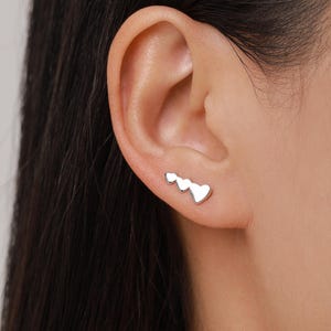 Può includere: Un orecchino rampicante in argento con tre disegni a forma di cuore. L'orecchino è indossato sull'orecchio, con i cuori che salgono di dimensione. Il gioiello è un accessorio semplice ed elegante.