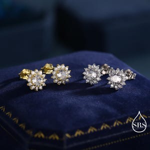 May include: Two pairs of stud earrings. One pair is gold-toned, the other is silver-toned. Each earring features a central, clear gemstone surrounded by smaller, clear gemstones, creating a floral or starburst design. The earrings are displayed on a dark blue velvet surface.