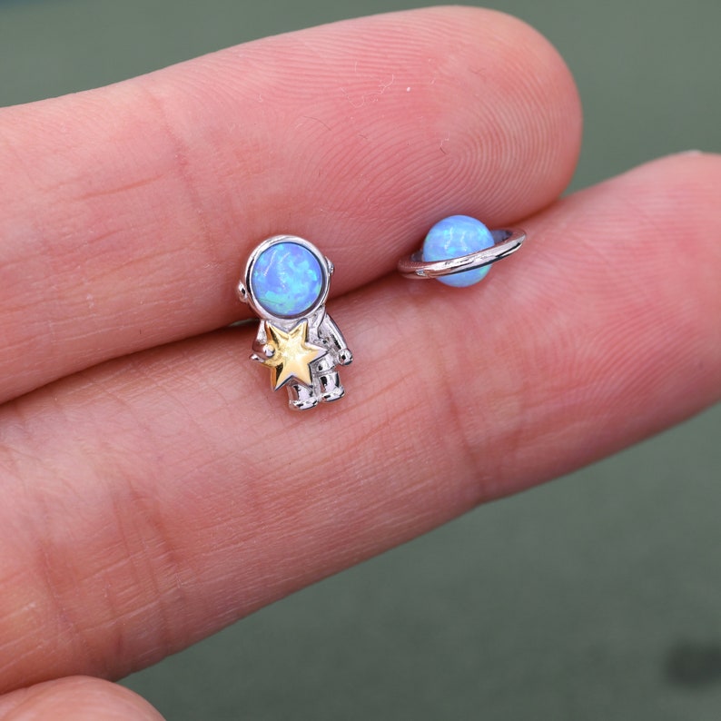 May include: A pair of silver-toned stud earrings. One earring is an astronaut figure holding a gold star. The other earring is a blue planet with a silver ring. Both earrings have a blue opal-like stone.