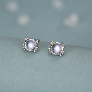 May include: A pair of silver-toned stud earrings shaped like cat heads. Each earring features a round, iridescent cabochon stone in the center, with small metal ears and three whiskers extending from the sides of the circular frame.