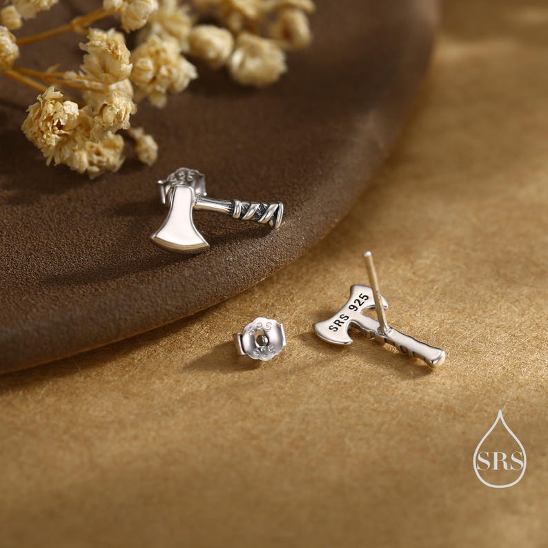 May include: A pair of silver axe-shaped stud earrings. The earrings have a detailed design with a white enamel axe head and textured handle. The earring post is visible on one earring, with "SRS 925" stamped on the axe head.